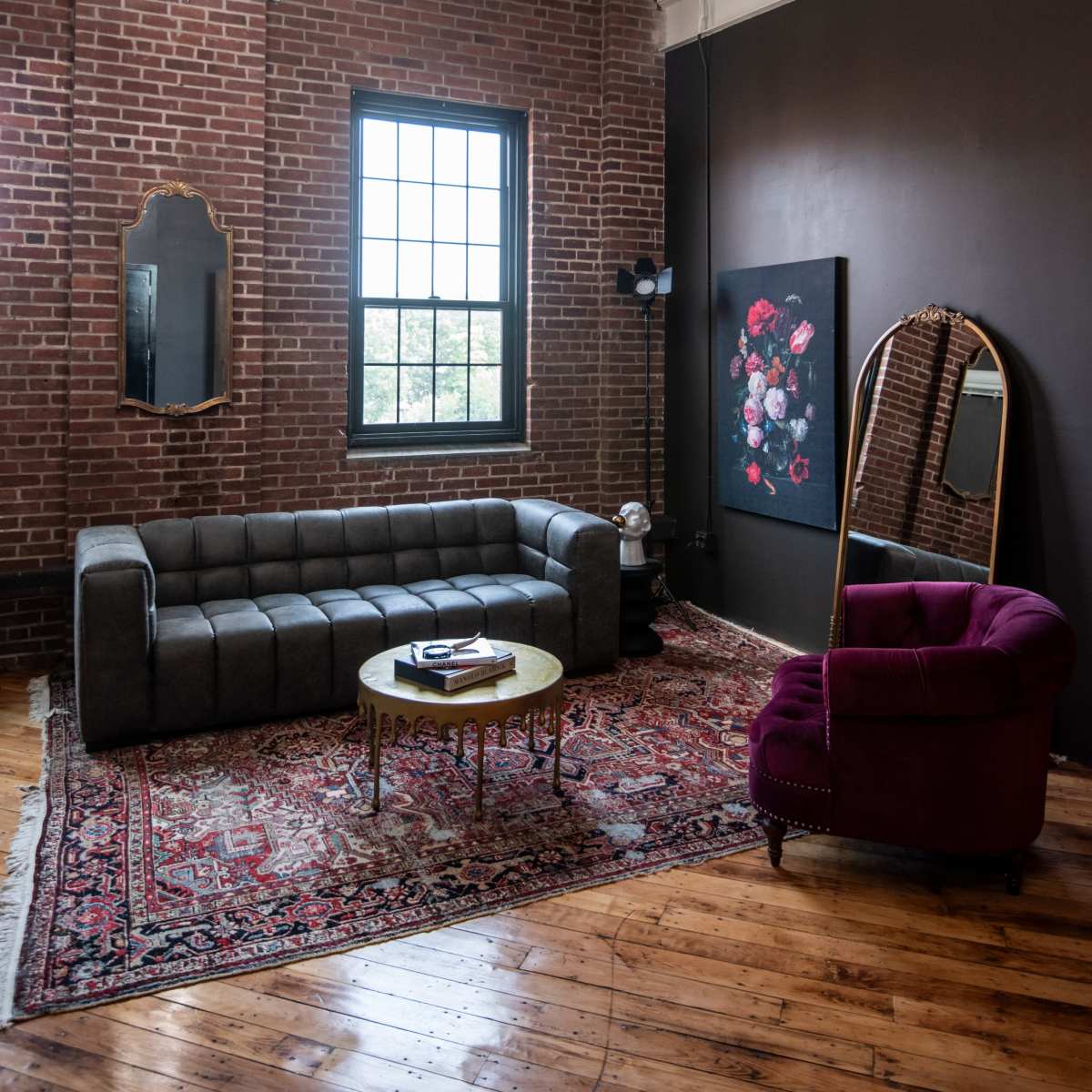 A living room with a brick wall, a large window, a gray sofa, a circular coffee table, a purple armchair, and a framed floral painting.