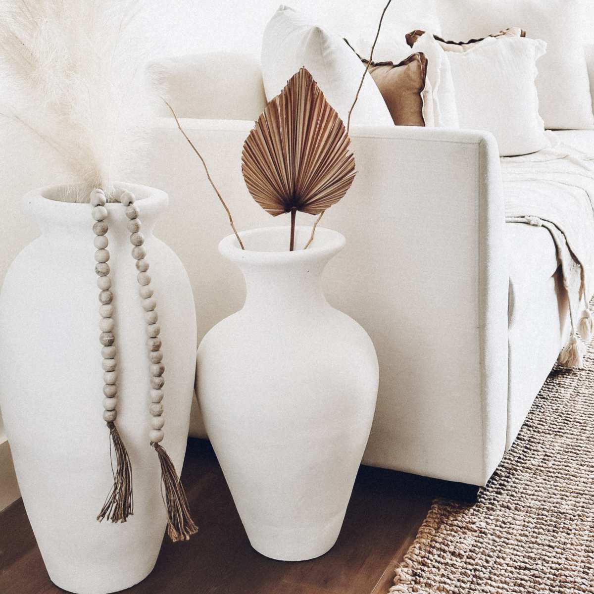 Two large white vases, one with decorative beads and the other holding a dried palm leaf, are positioned beside a light-colored sofa in a bright living room.