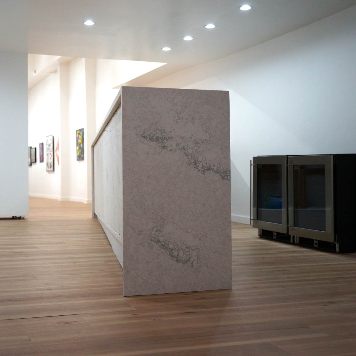 A modern interior space with light wood flooring, a large white counter or island, and a wall adorned with framed artwork in the background.