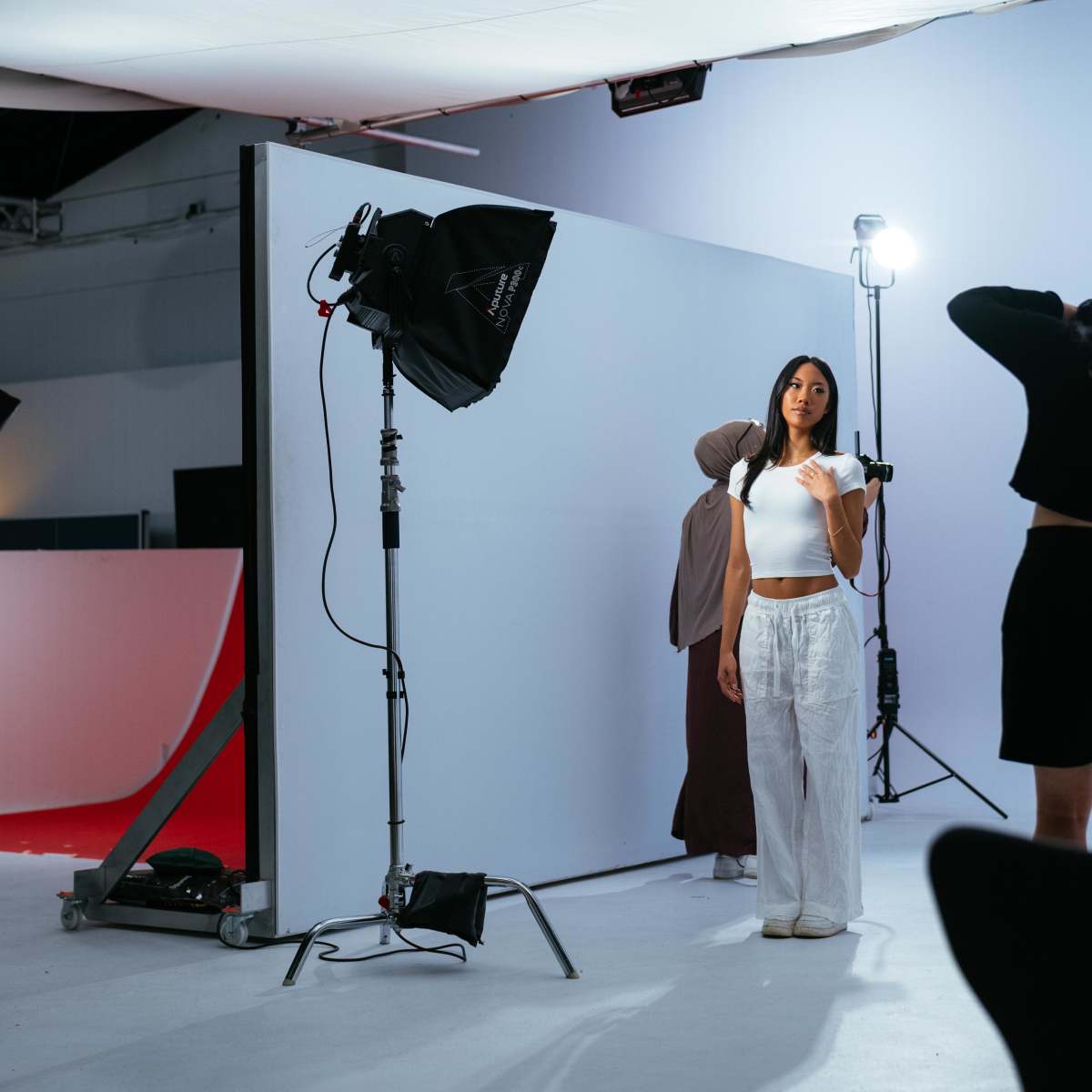 A fashion photoshoot is taking place in a studio with a model posing against a white backdrop.