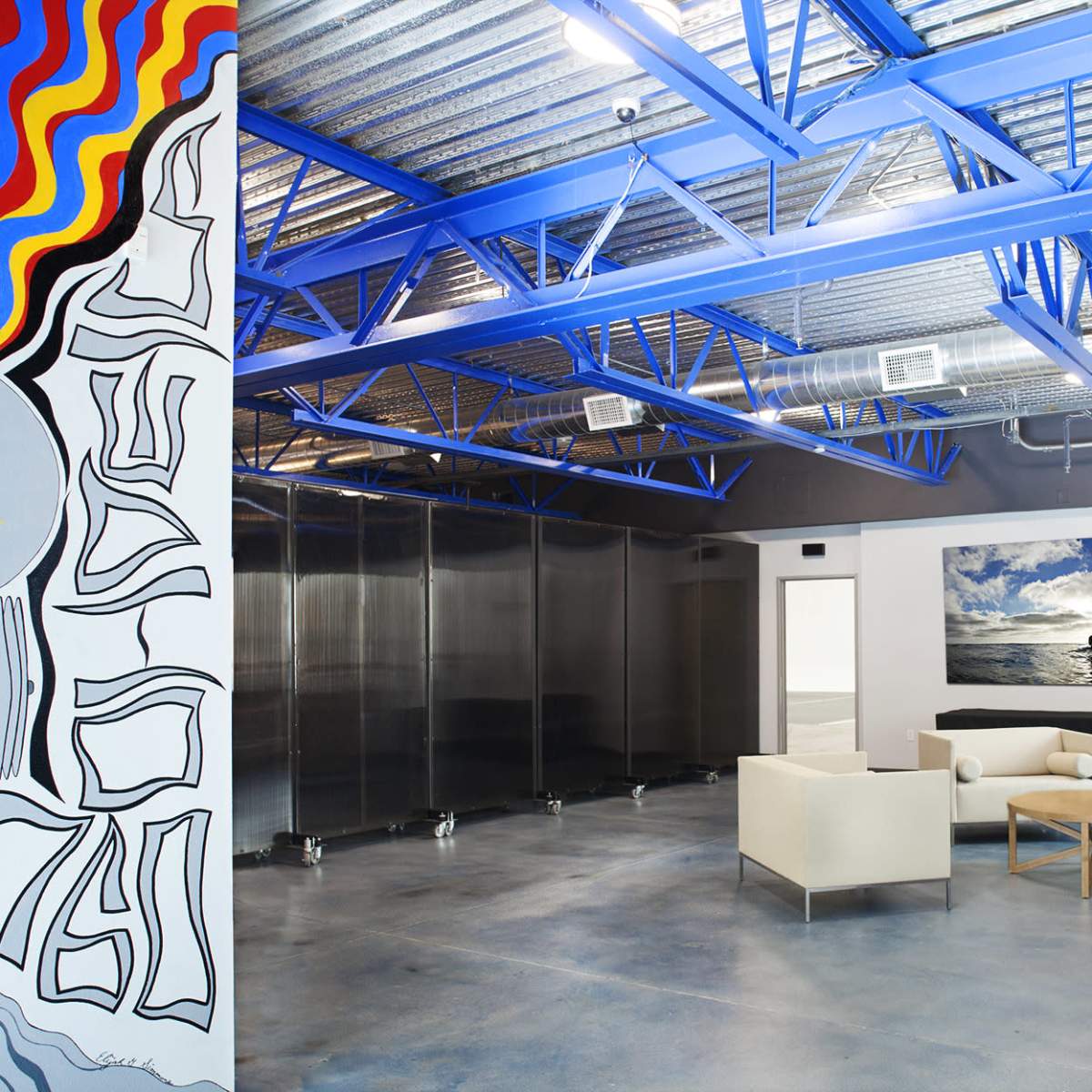 A modern office space with a colorful mural on the left, large glass partitions, and a seating area with two sofas and a coffee table.