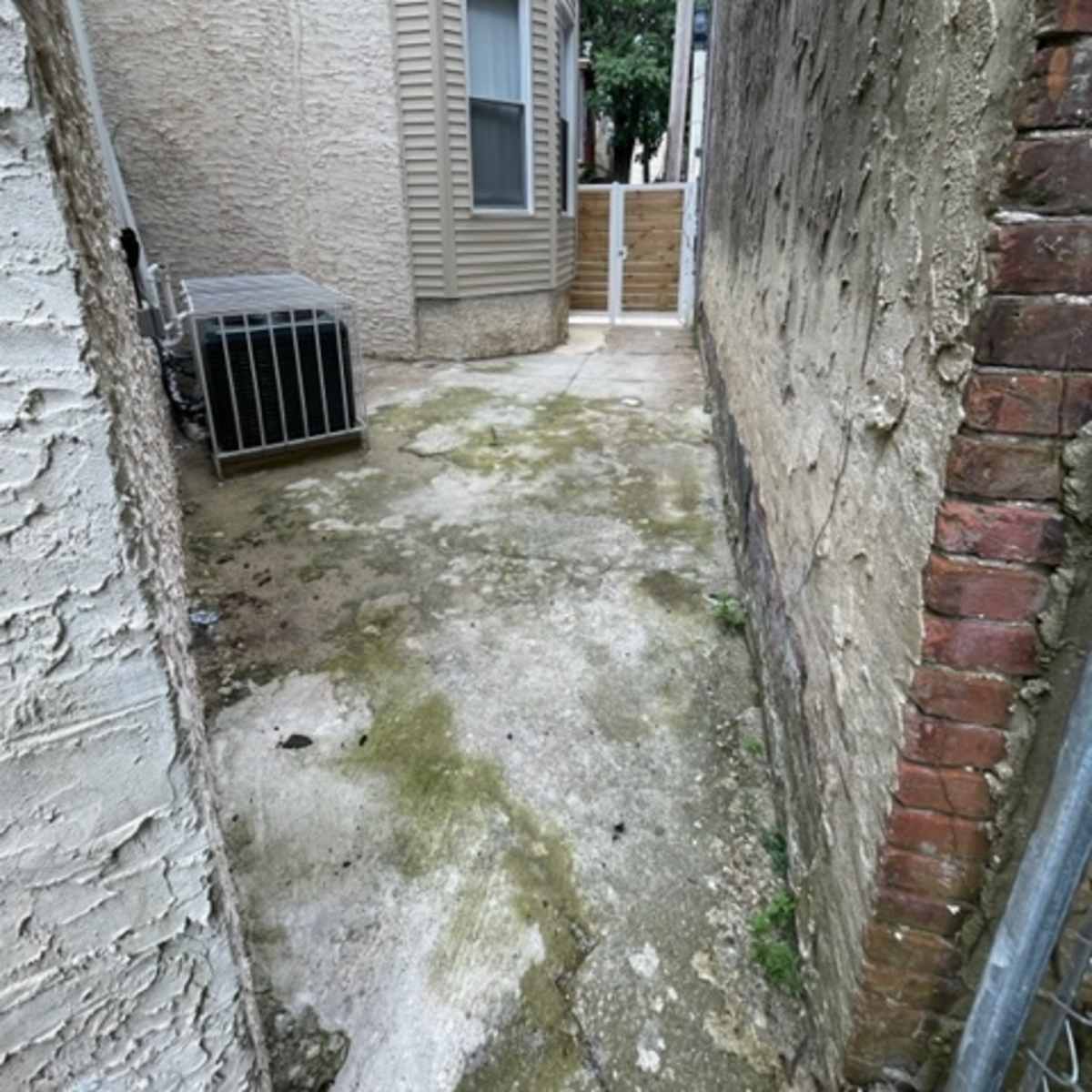 A narrow alleyway between two buildings, with a concrete surface strewn with debris and an air conditioning unit visible on the left.
