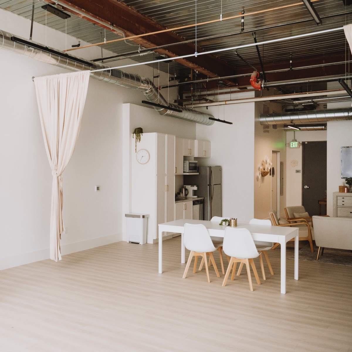 A minimalistic, bright interior space featuring a white kitchen setup, a dining table with chairs, and large windows, with a curtain partially drawn.