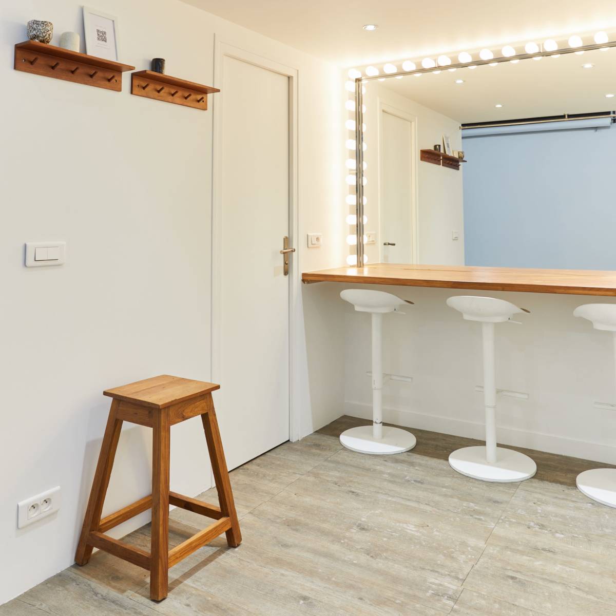 A modern room featuring a wooden bar with three white stools, illuminated by a lighted mirror and a shelf mounted on the wall.