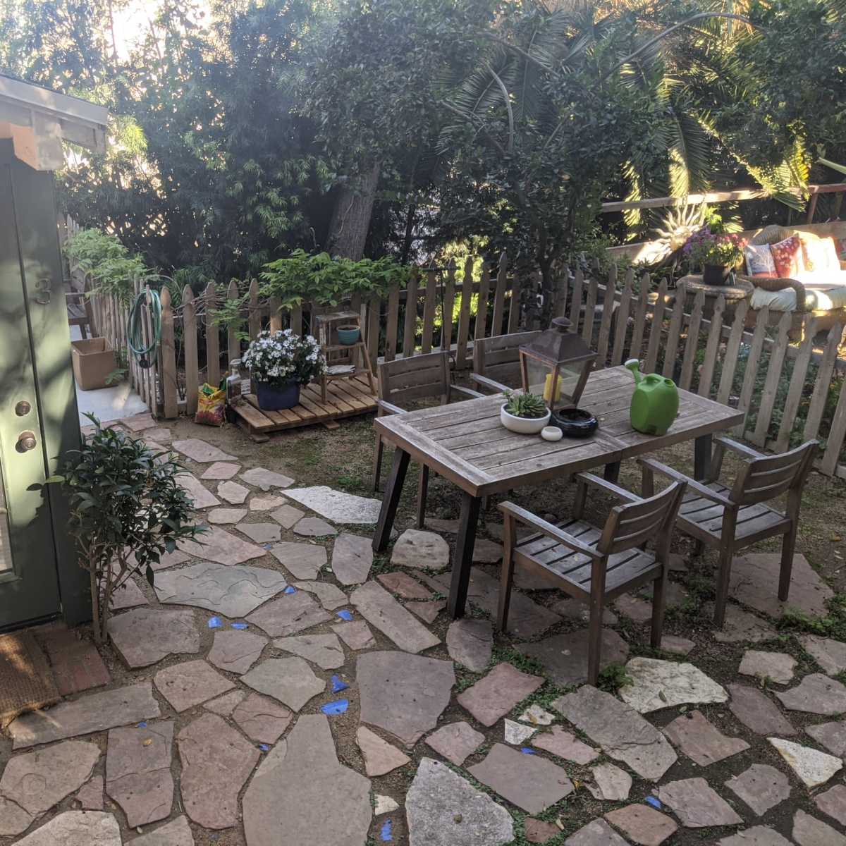 A backyard with a stone pathway, a wooden table and chairs, a small green building, and various plants surrounded by a wooden fence.