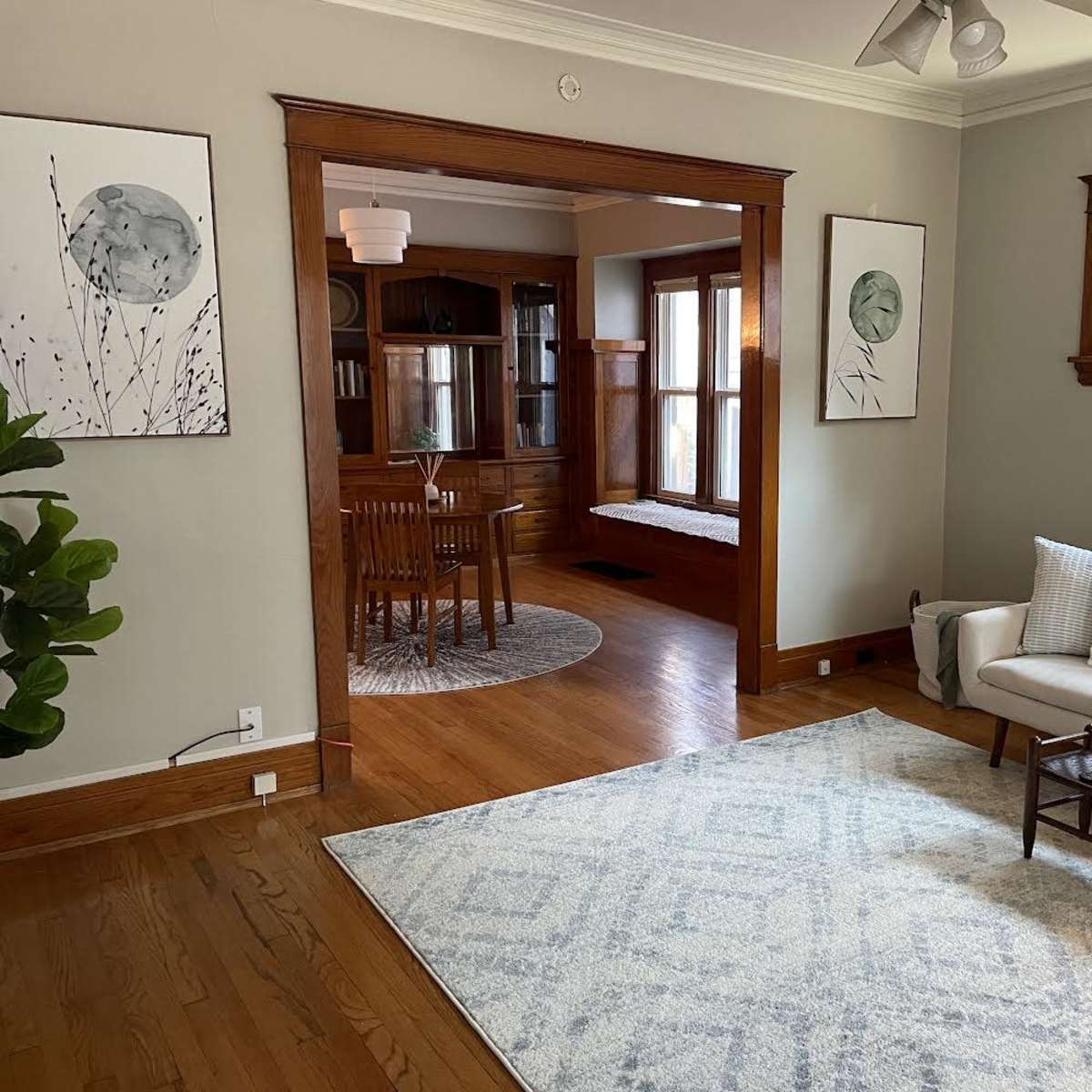 A light-filled living room features a beige sofa, a decorative rug, and two framed botanical prints on the walls, with an adjoining dining area visible through an archway.