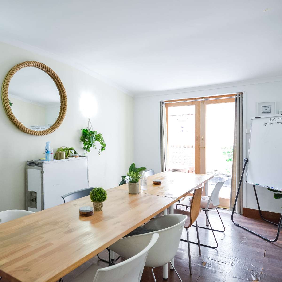 A bright meeting room features a wooden table surrounded by chairs, a mirror on the wall, and a whiteboard near a window.