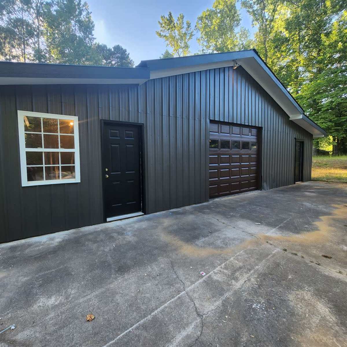 A dark-colored garage with a single door and a window, set against a backdrop of greenery.