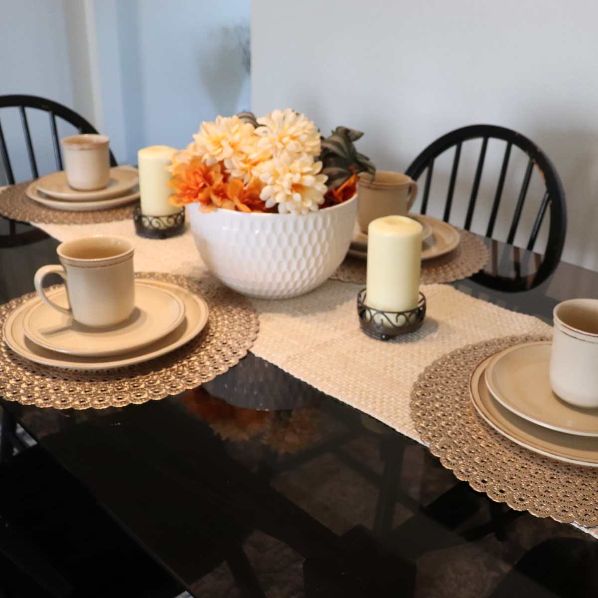 A dining table is set with beige plates and mugs, adorned with a floral centerpiece and lit candles on a runner.