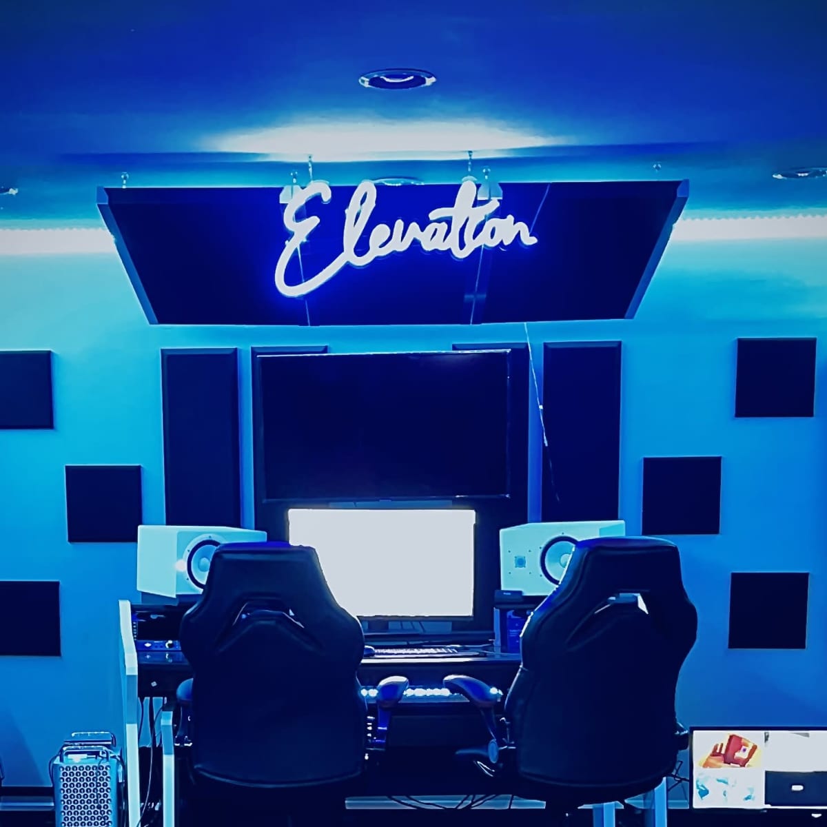 A modern recording studio with two black office chairs facing a computer setup beneath a blue neon sign that says "Elevator."