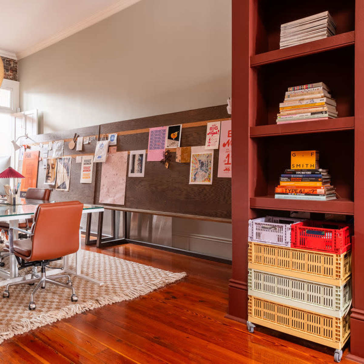 A modern office space features several glass desks with ergonomic chairs, a wooden accent wall displaying various artworks, and a red bookshelf filled with colorful baskets and books.