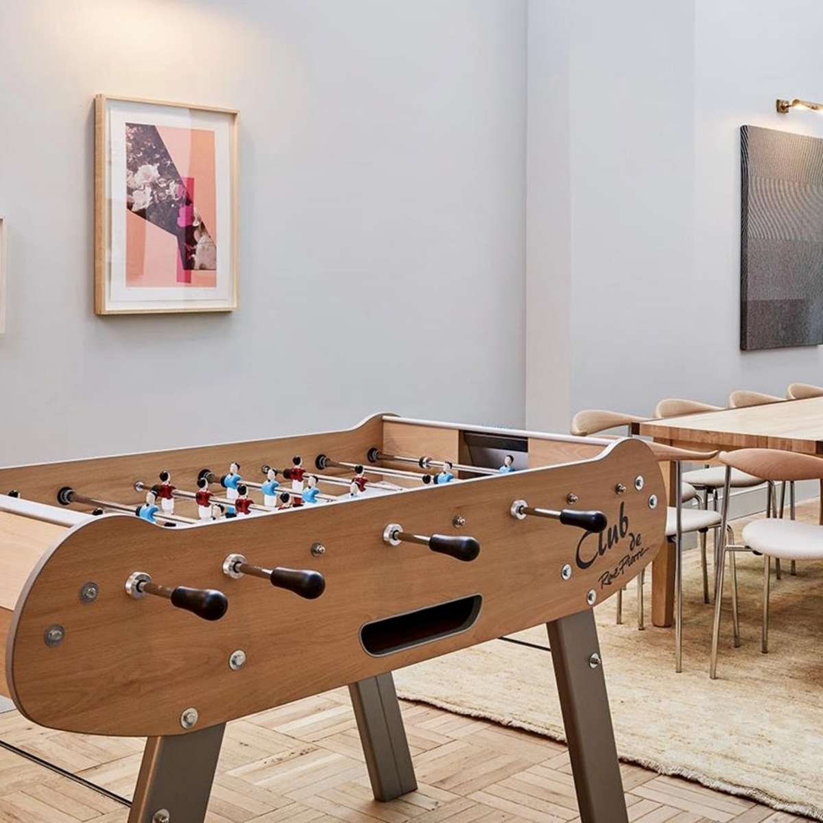 A spacious room with a foosball table in the foreground and a long dining table surrounded by chairs in the background.