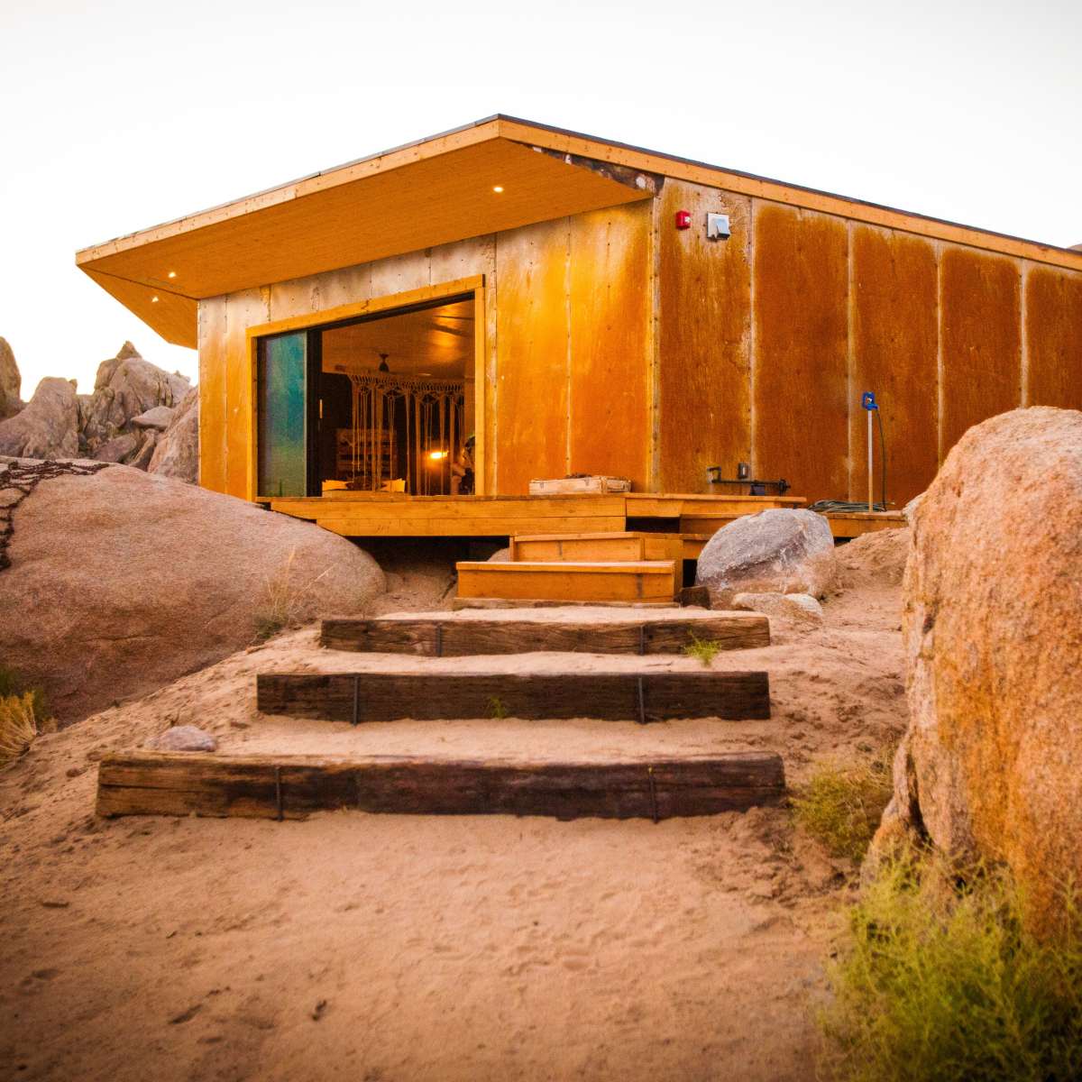 A modern cabin is set against a backdrop of large boulders with wooden steps leading up to its entrance.