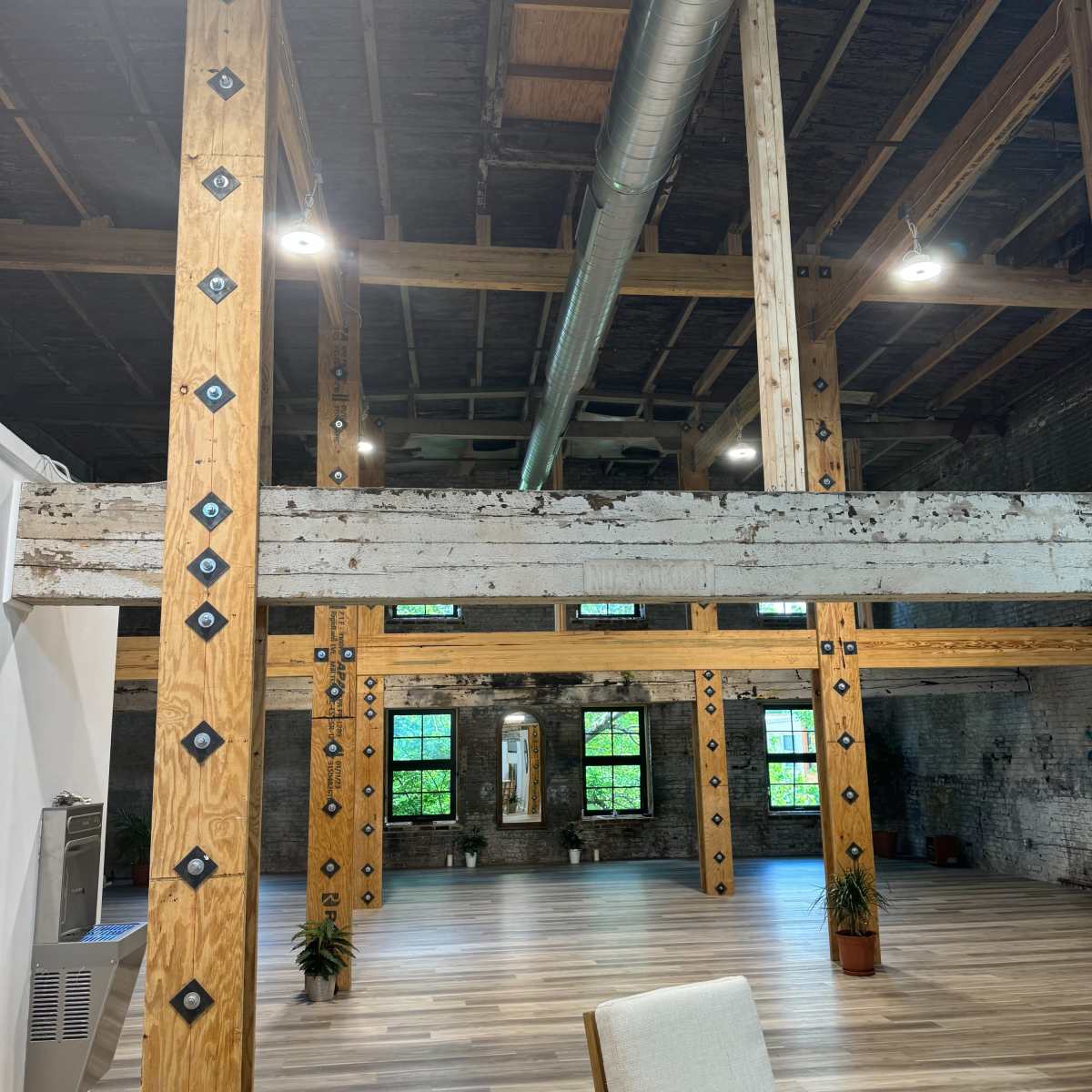 An industrial-style interior space with exposed wooden beams, metal accents, and large windows overlooking a wooden floor.