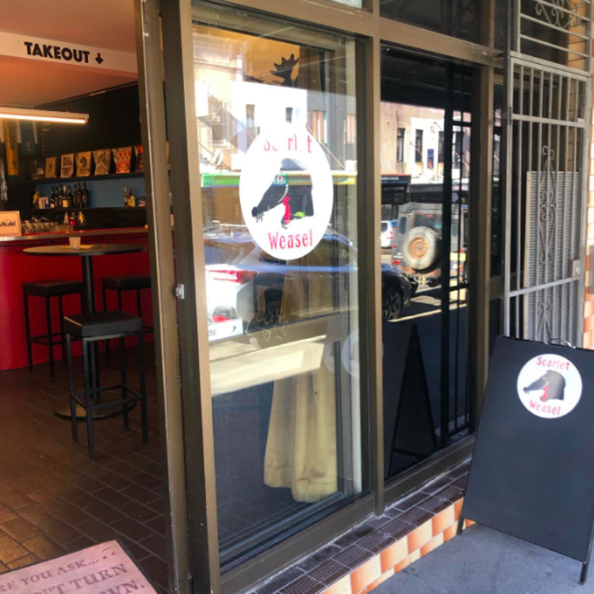 The entrance of a restaurant called "Scarlet Weasel," with a logo displayed on the window and seating visible inside.