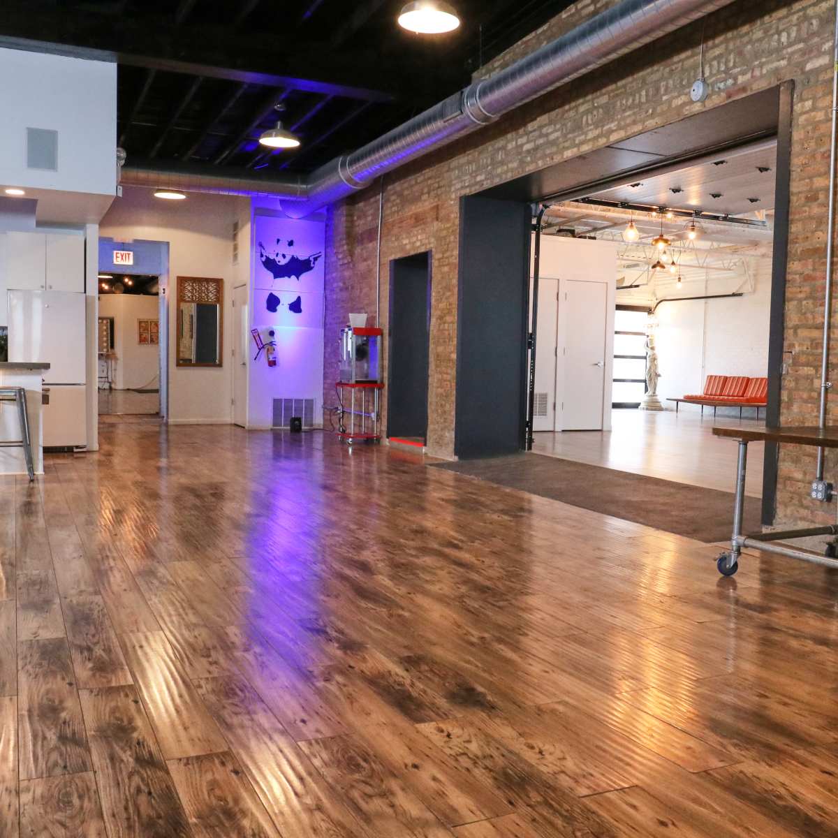 A spacious industrial-style room with wooden floors, exposed brick walls, and a kitchen area on one side.