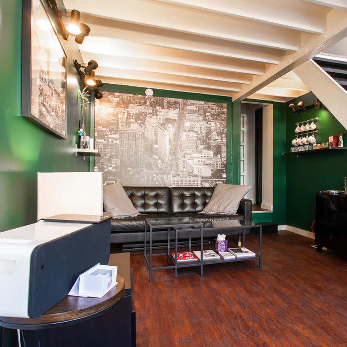 A small living area features a black leather couch, a wall with a cityscape mural, a staircase, and a coffee station.