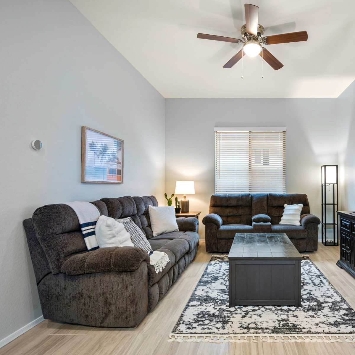 A living room with two sofas, a coffee table, and a large television mounted on the wall, complemented by a ceiling fan and a window with blinds.