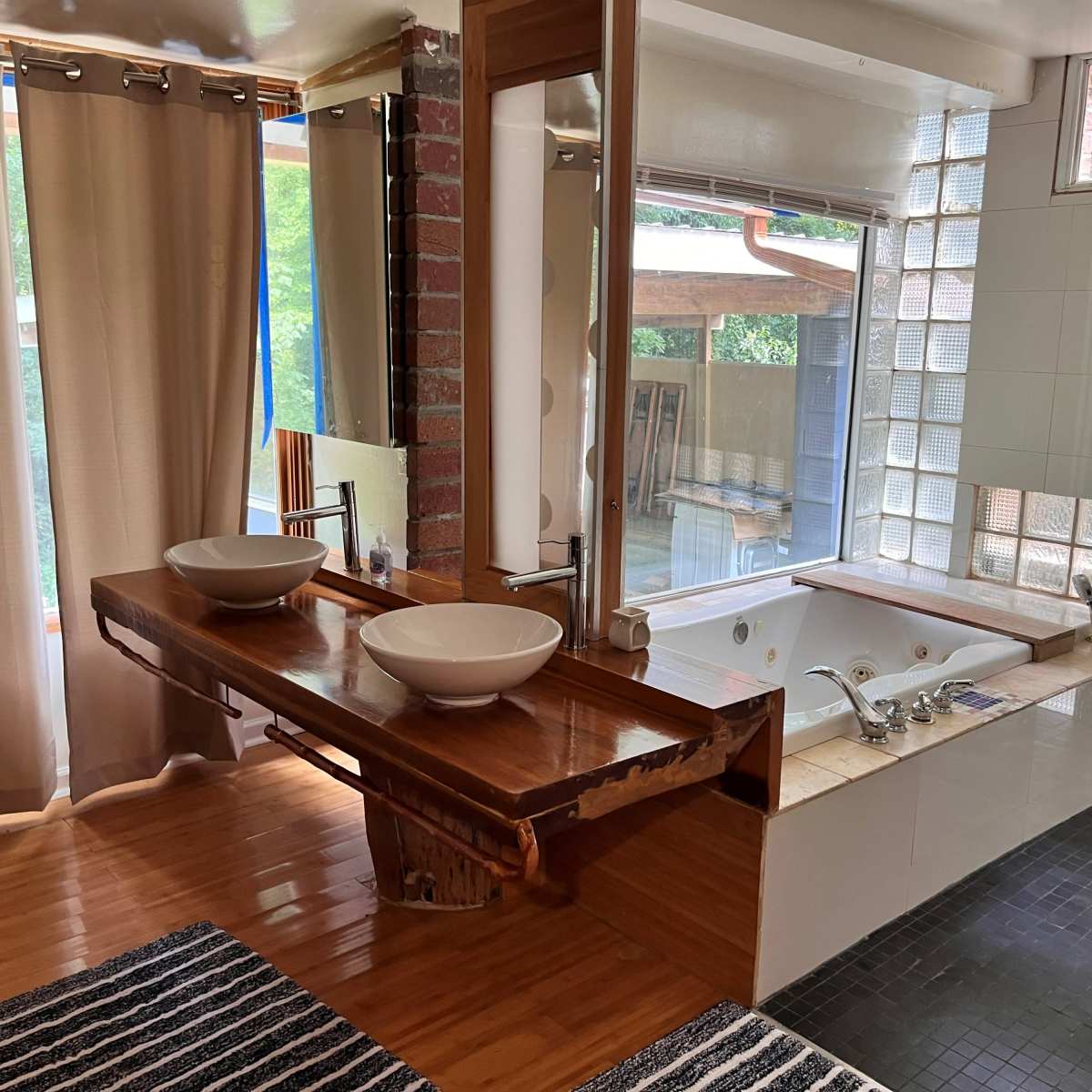 A modern bathroom featuring a wooden vanity with two sinks, large mirrors, and a spacious area with a jacuzzi tub and a glass block wall.