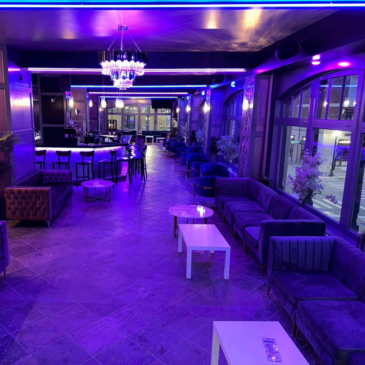 A modern lounge with an illuminated bar area and stylish seating, featuring purple lighting and decorative plants.