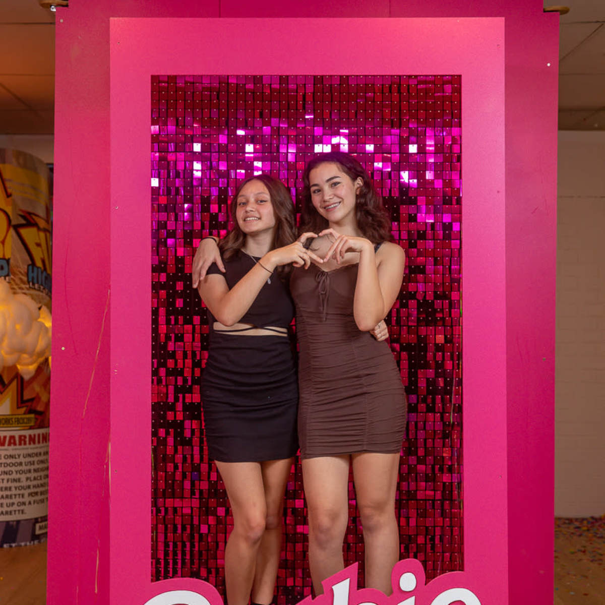 Two young women stand together in a brightly decorated frame with a pink background and "Barbie" written in white at the bottom, creating a playful photo setting.
