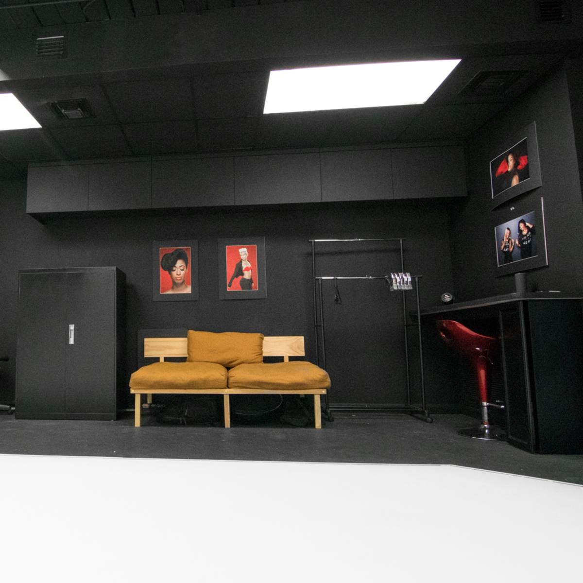 A photography studio with black walls, a white backdrop, a wooden sofa, and framed artworks.