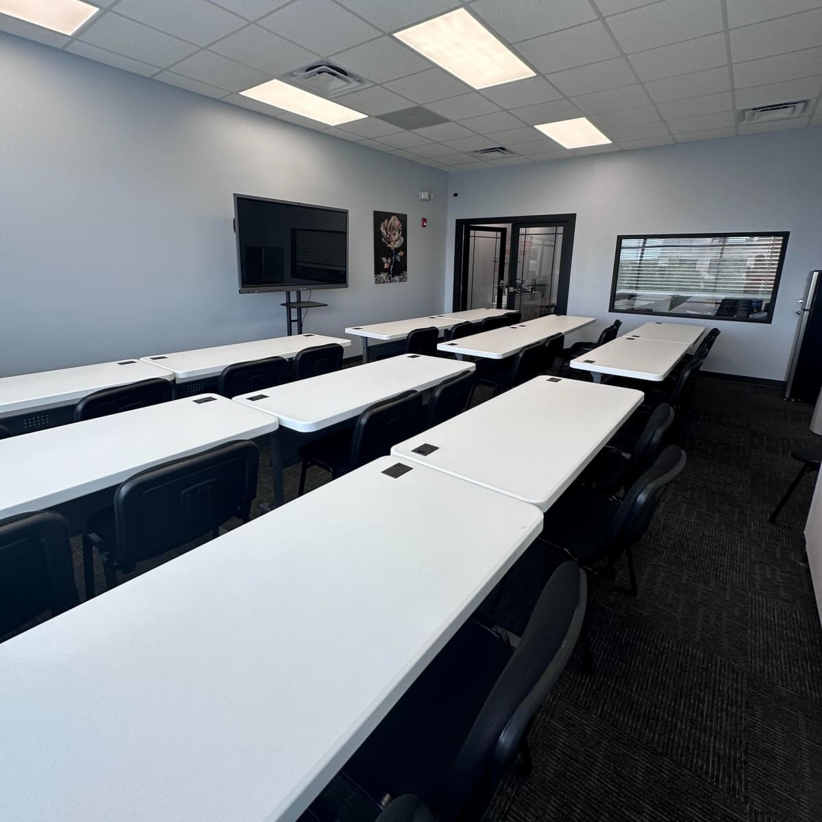 A modern classroom setup with multiple white tables arranged in rows and a large screen mounted on the wall.