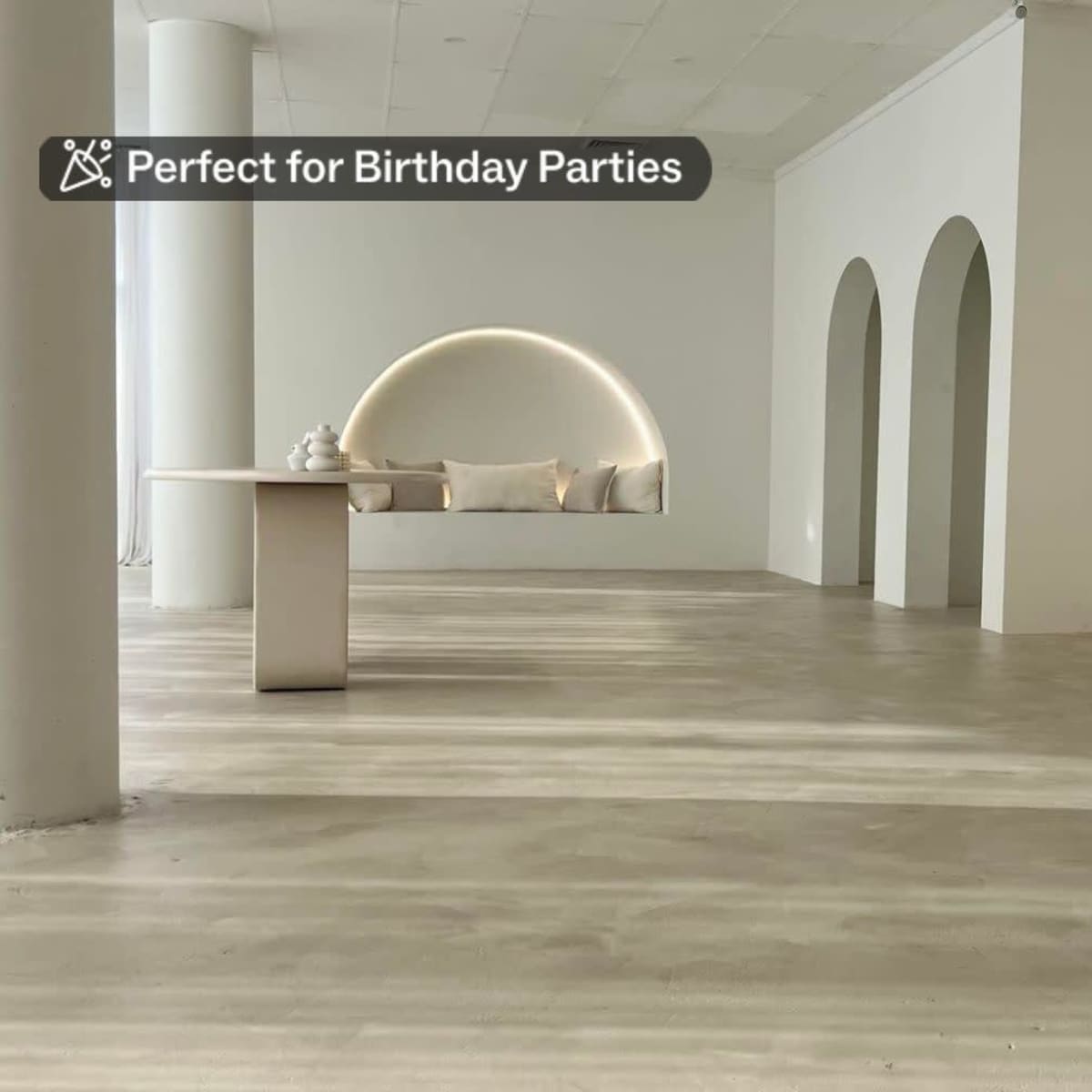 A minimalistic interior space with a large mirror, a simple table, and archways on the walls, featuring a light-colored floor and soft seating.
