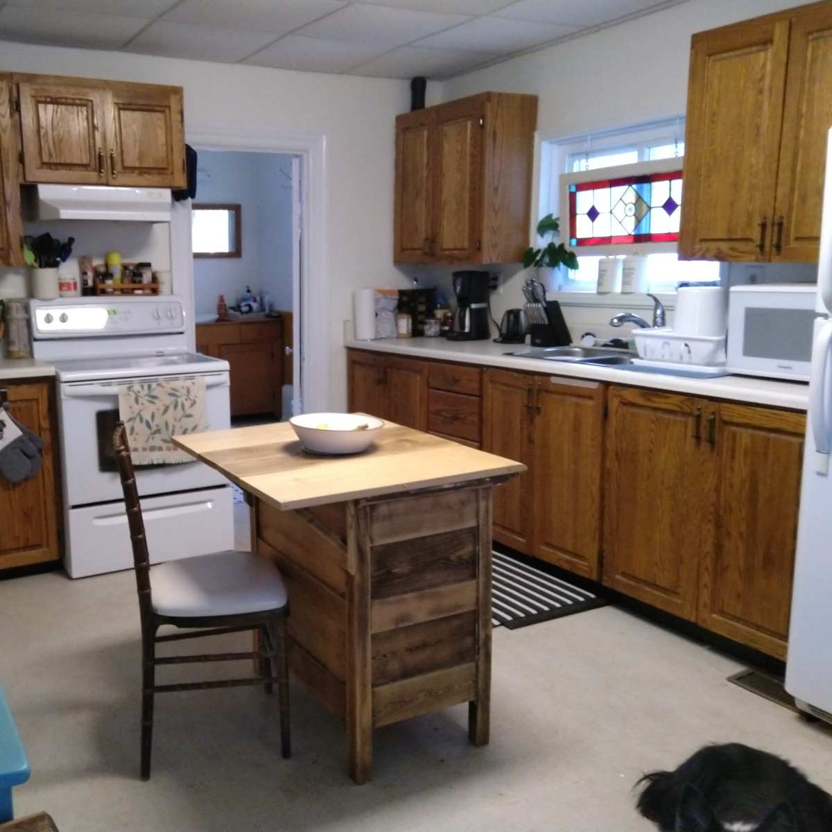 A compact kitchen features wooden cabinets, a small dining table with a bowl, and various appliances along the countertops.