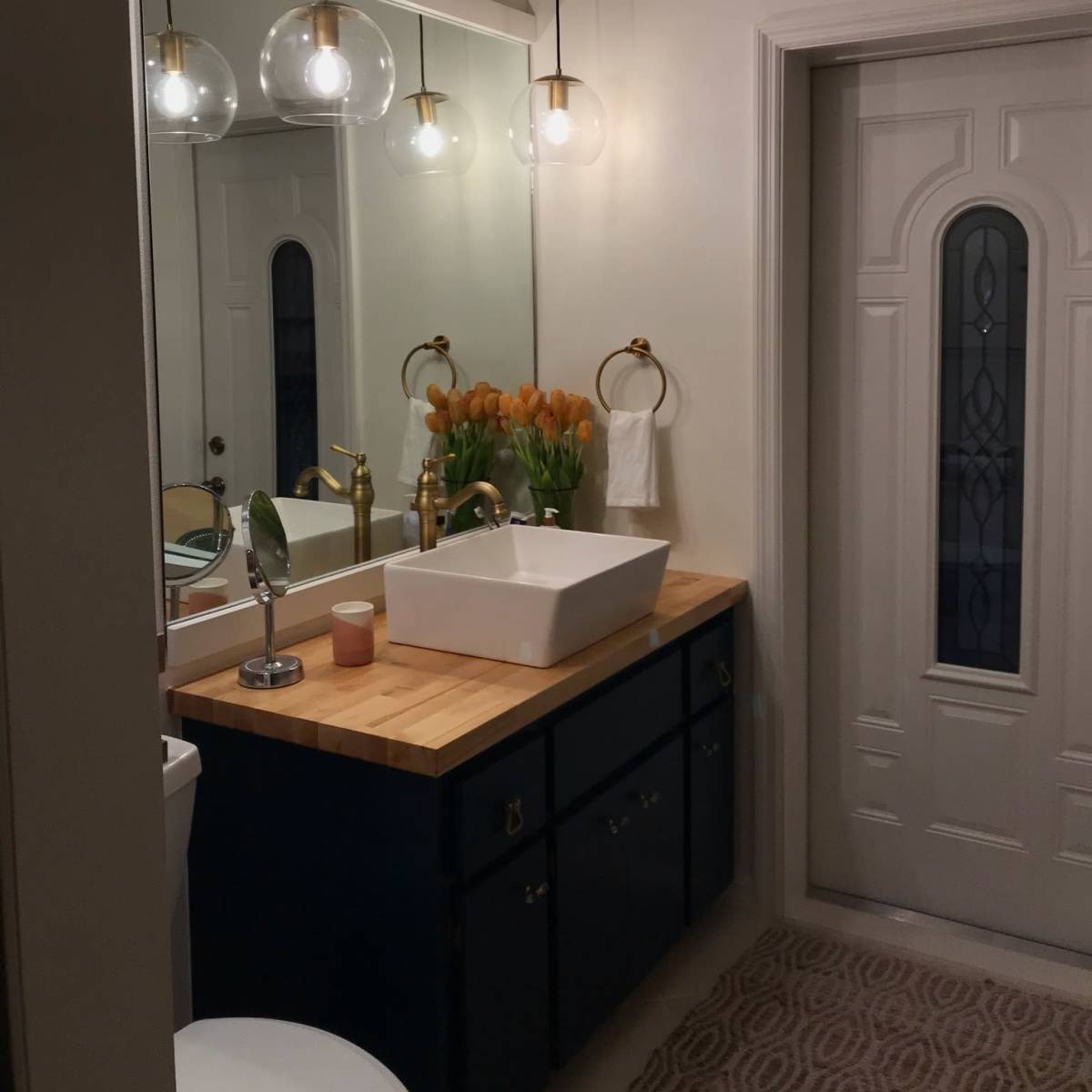 A modern bathroom with a wooden countertop, a white rectangular sink, a large mirror, and a vase of orange tulips, with a door leading to another room.