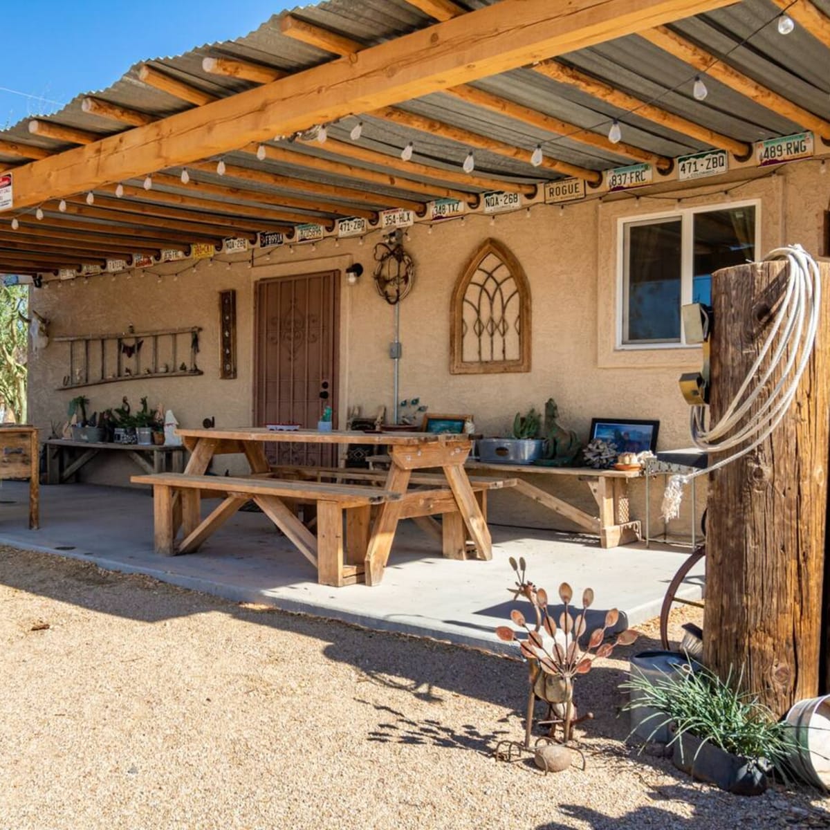 A rustic outdoor seating area with a wooden picnic table under a covered patio, surrounded by various decorative items and plants.