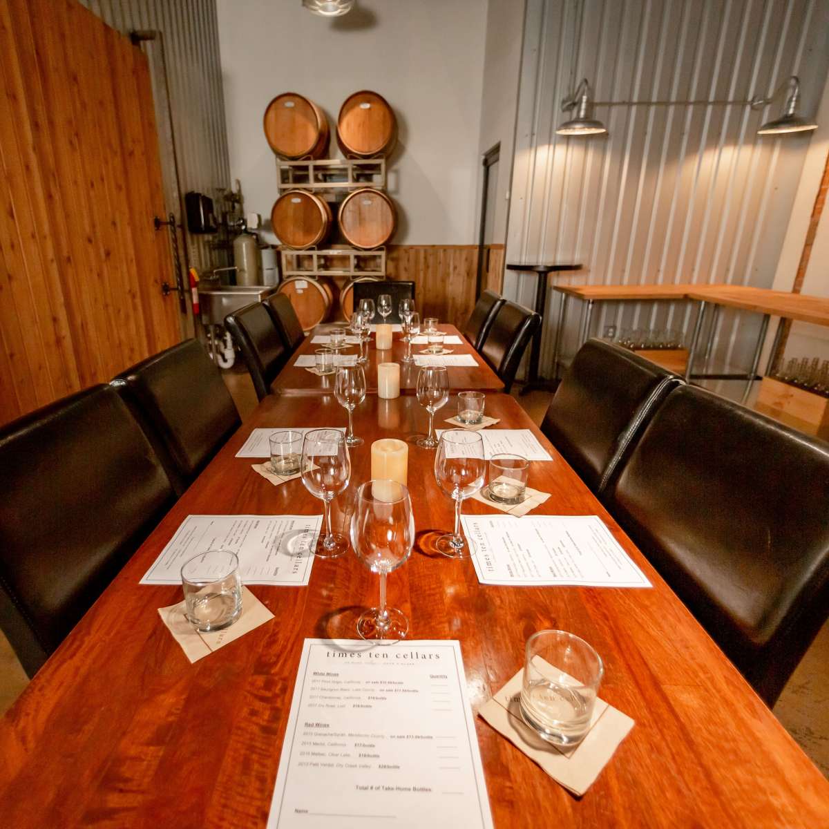 A long wooden dining table is set with glasses and menus in a room featuring wooden barrels and metal walls.