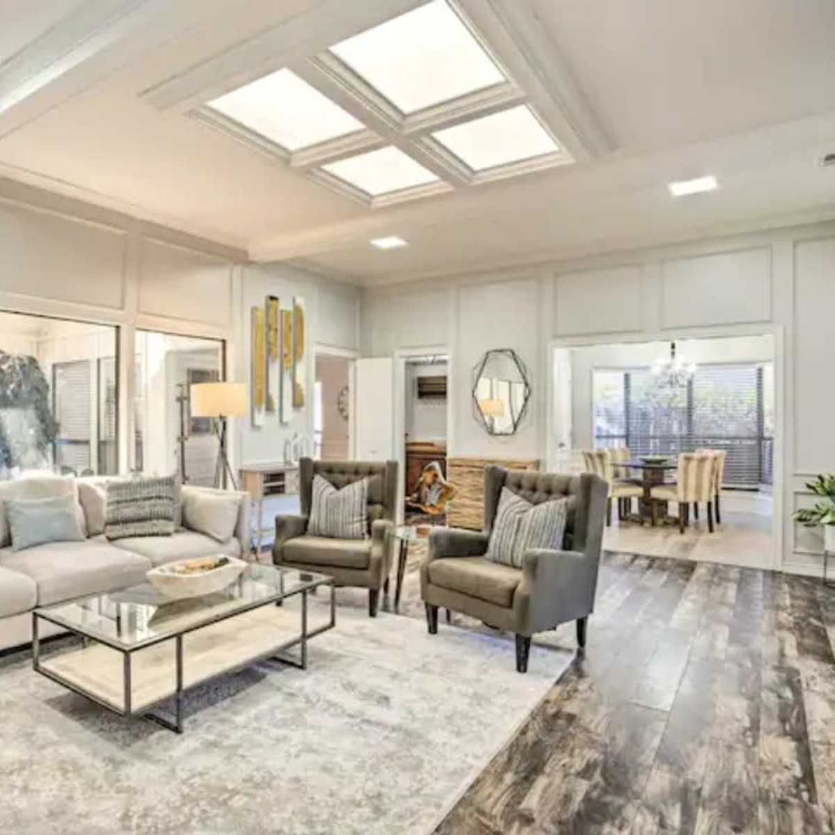 The living room features a light-colored sofa, two armchairs, a glass coffee table, and large windows that overlook a patio area.