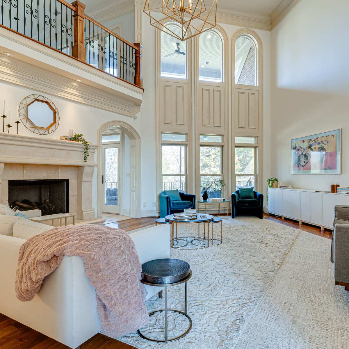 A spacious living room with high ceilings, featuring a stone fireplace, large windows, and modern furniture arranged around a central area rug.