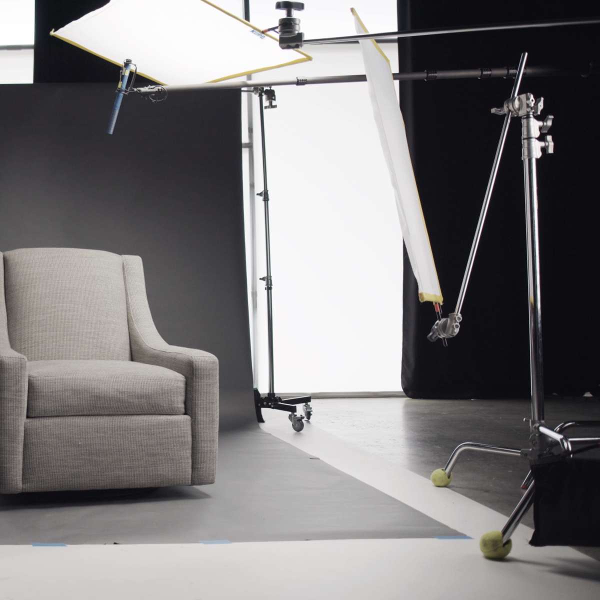 A gray armchair is positioned on a black floor mat in a studio setting with backdrop lighting and equipment.