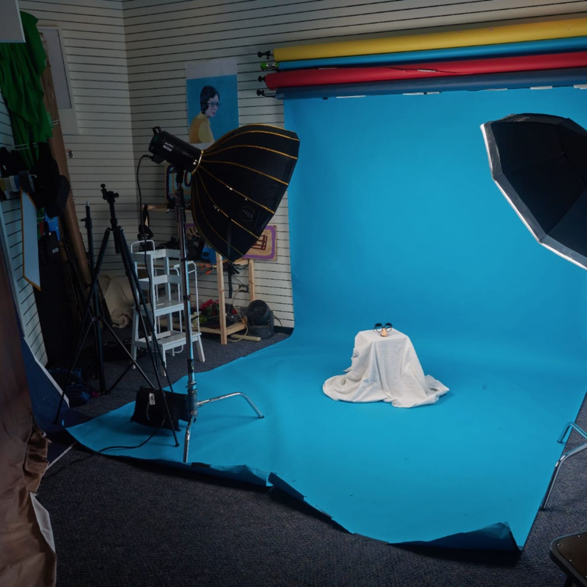 A photography studio with a blue backdrop, two softbox lights, a table covered with a white cloth, and various photography equipment arranged around the space.