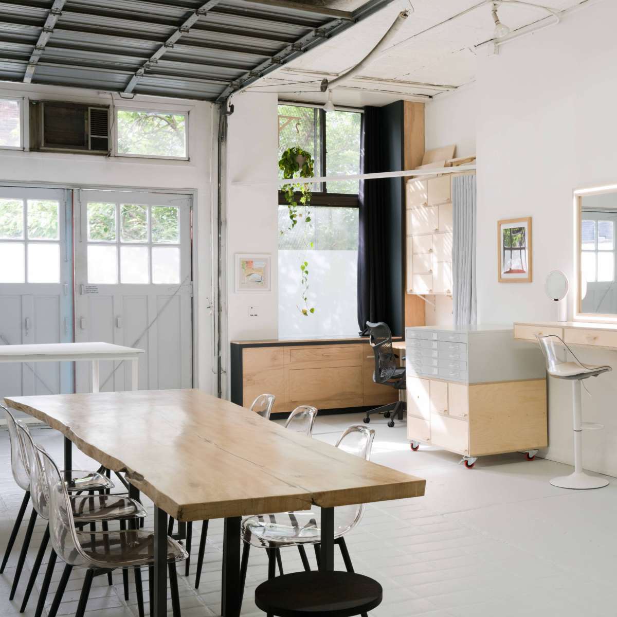 A spacious, modern workspace features a long wooden table, clear acrylic chairs, and mirrors on a wooden vanity against white walls.