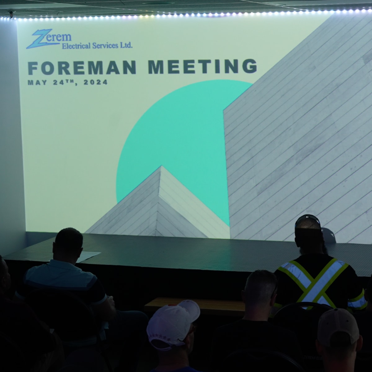 A presenter stands in front of a screen displaying "Foreman Meeting" along with the date May 24th, 2024, while an audience seated in semi-darkness listens attentively.