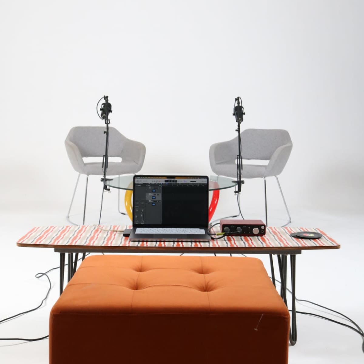 A minimalist studio setup with two microphones, two chairs, a laptop, and an orange ottoman on a striped table.