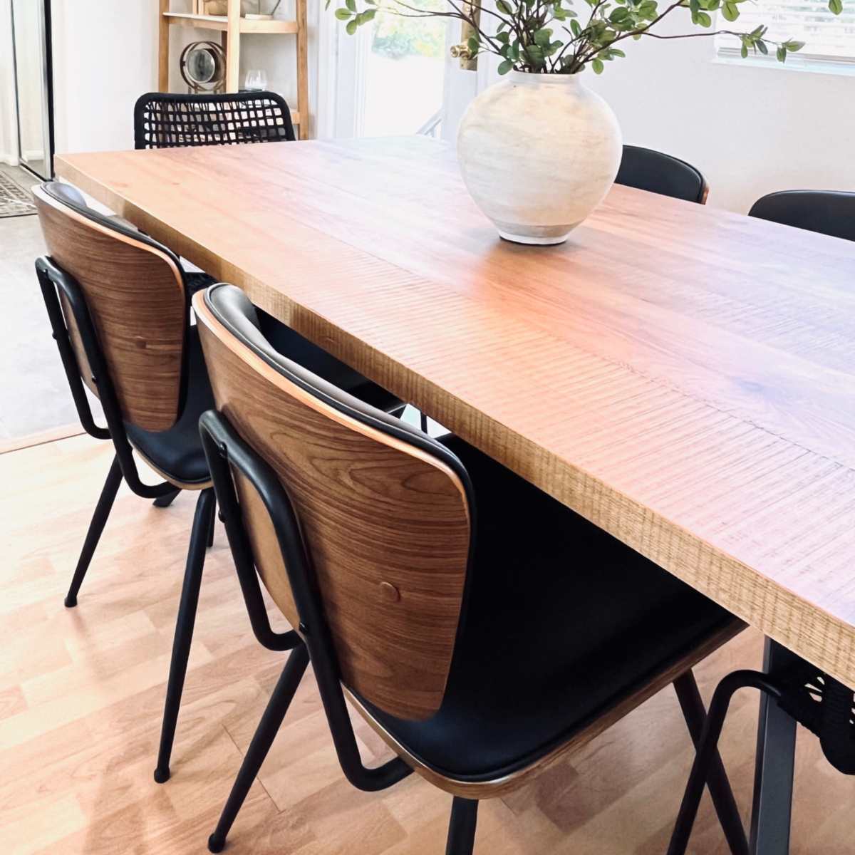 A wooden dining table is set with a vase of greenery, surrounded by black chairs in a well-lit room.