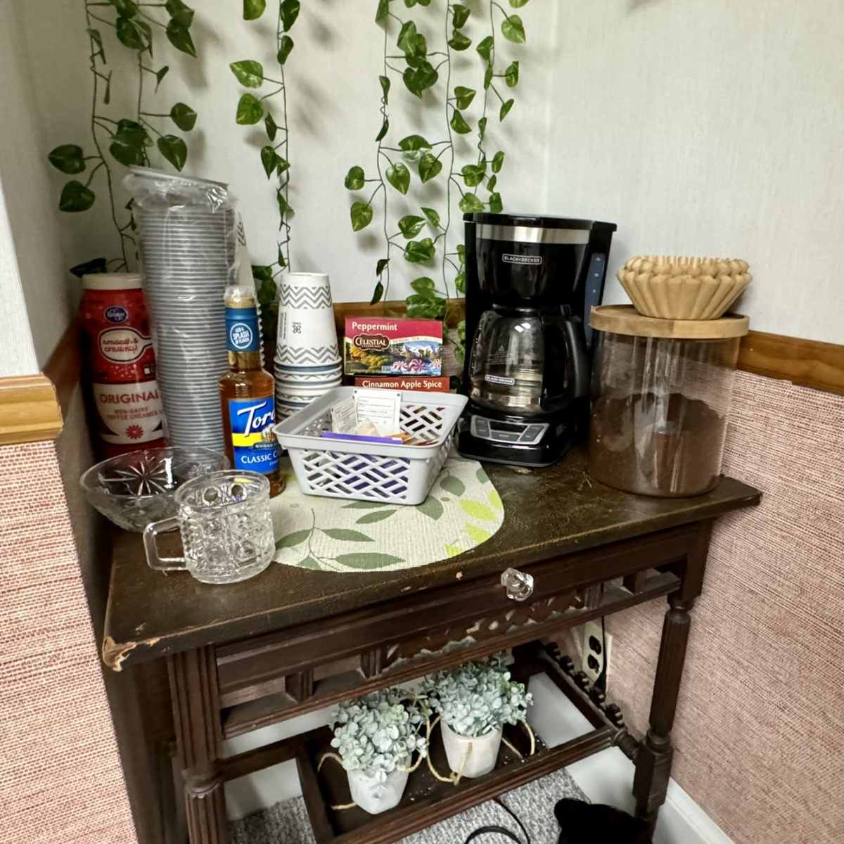 A corner table with a coffee maker, several containers, and glasses, surrounded by green plants on the wall.