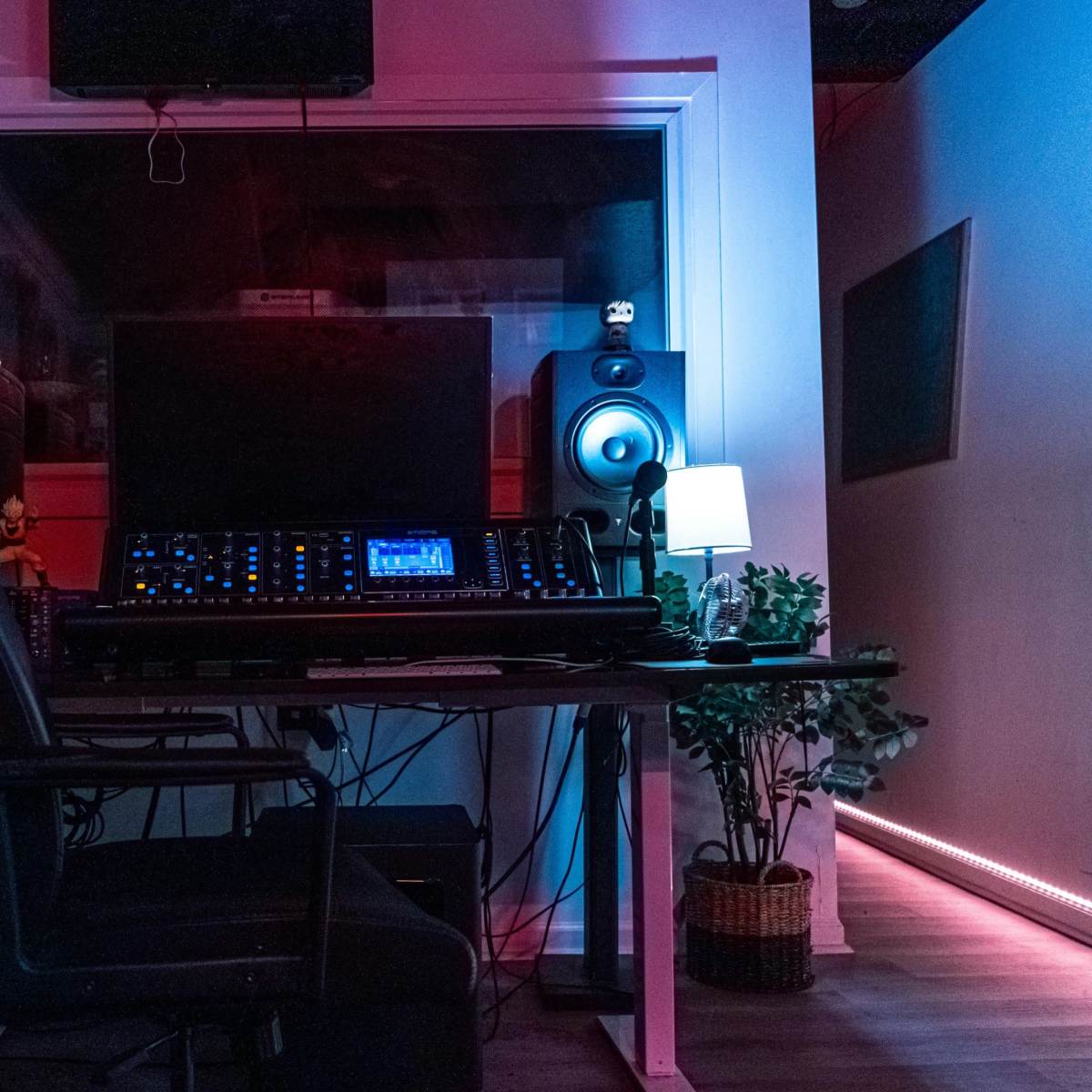A music studio with a desk featuring audio equipment, a chair, and a small lamp, illuminated by colored lights.
