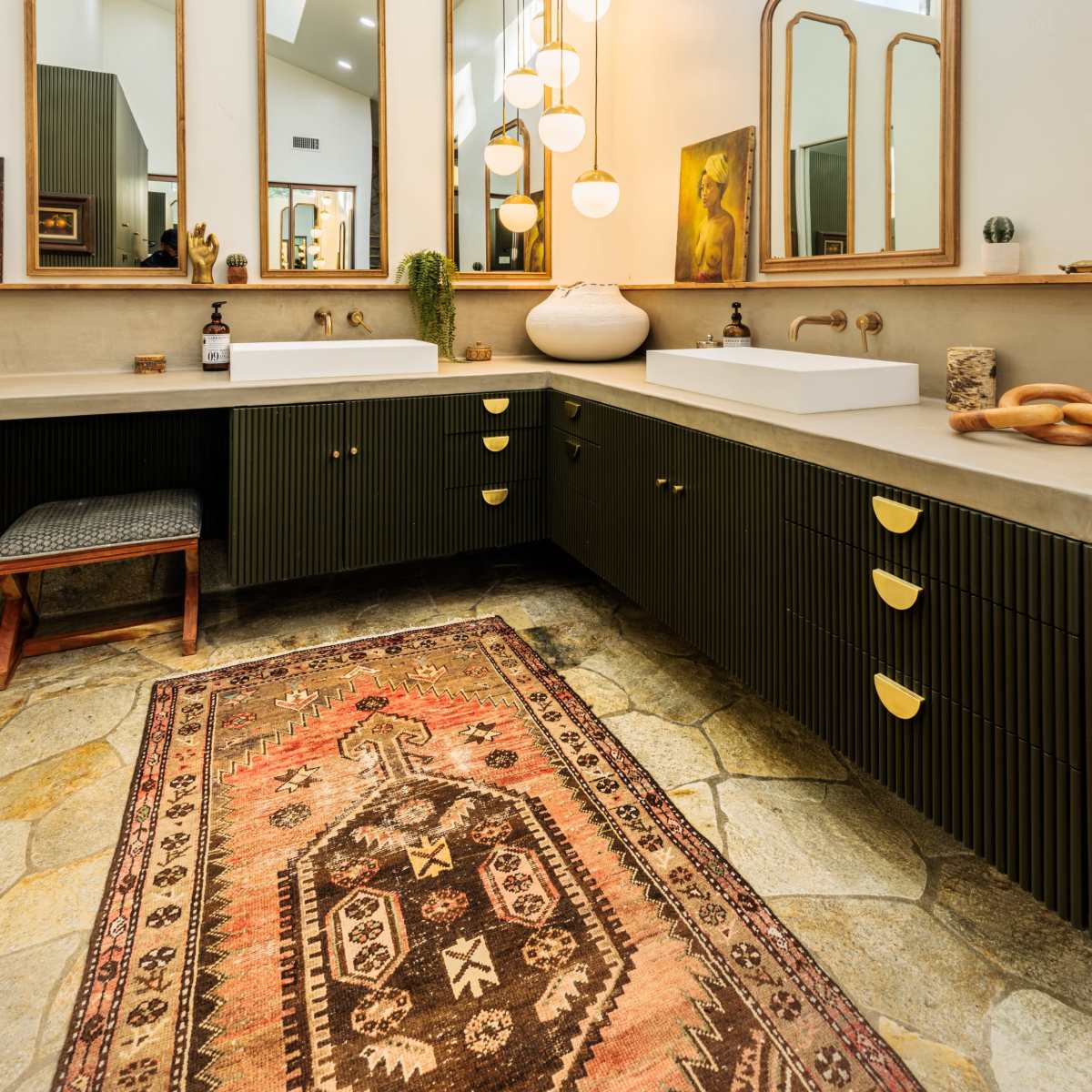 The bathroom features a double vanity with green cabinets, multiple mirrors, a light fixture hanging above, and a patterned rug on a stone floor.