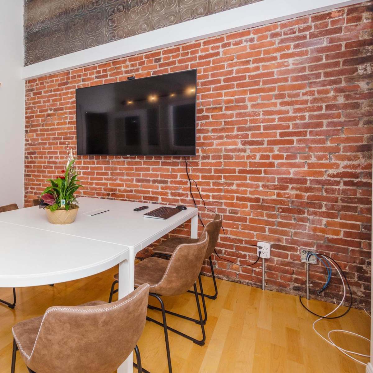 A conference room with a white table, brown chairs, a plant centerpiece, and a wall-mounted television on a brick wall.