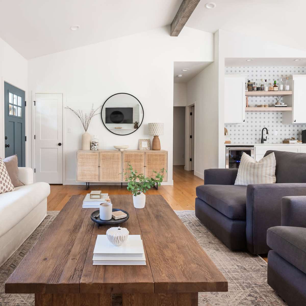 A modern living room features a white sofa and dark armchair around a wooden coffee table, with a large mirror and a sideboard on the walls.