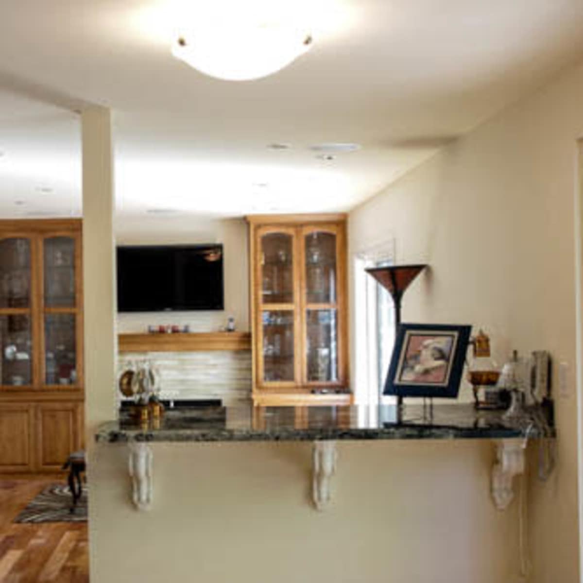 A well-lit interior space features a kitchen counter with a granite surface facing a living area that includes wooden cabinetry and a mounted television.