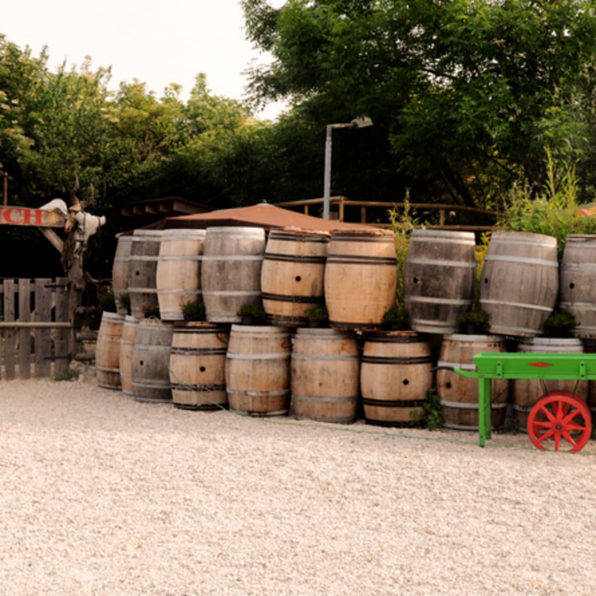 A stack of wooden barrels is positioned next to a green cart in front of a ranch entrance surrounded by trees.