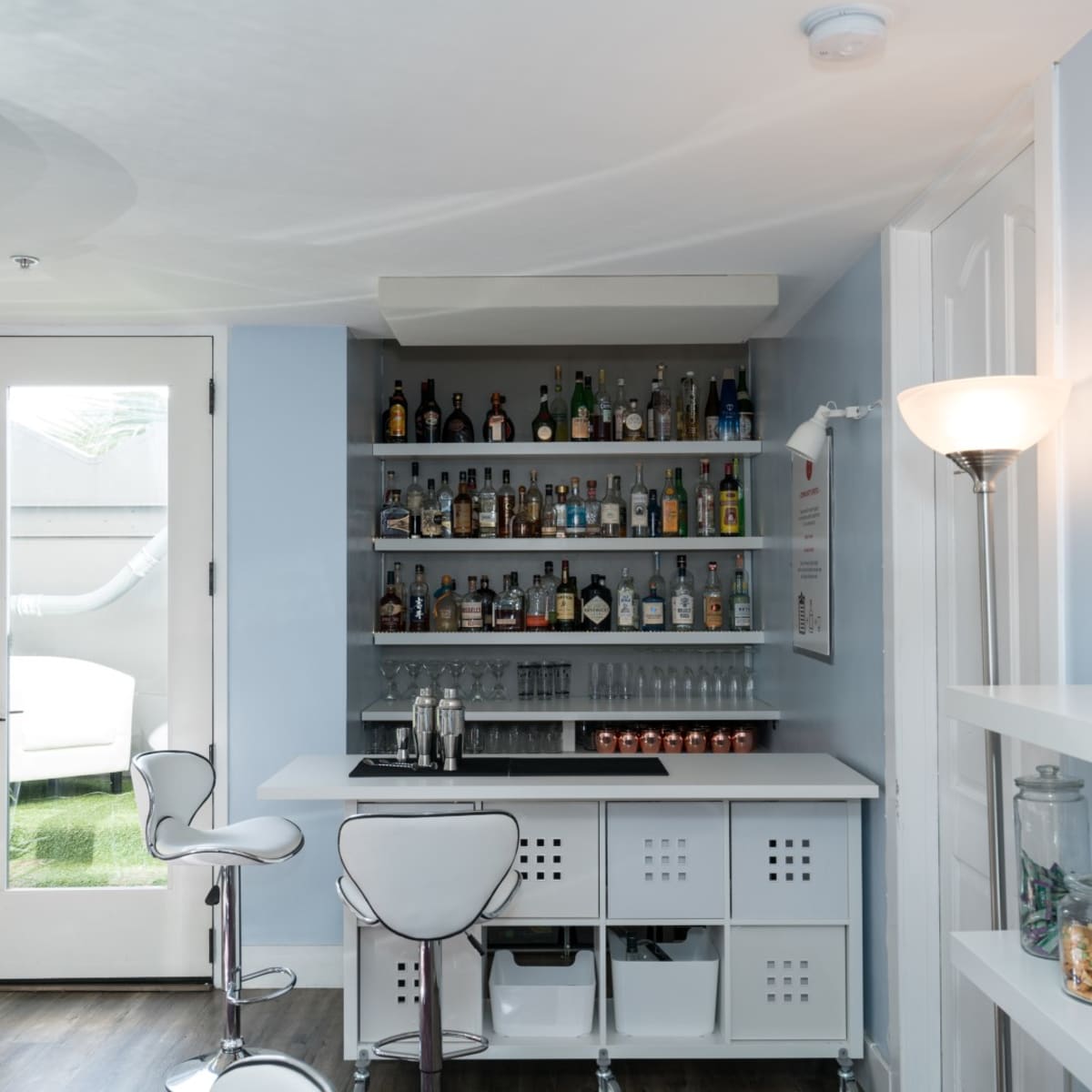 A modern bar area with a white counter, high stools, and shelves lined with various bottles, alongside a glass door leading to an outdoor space.