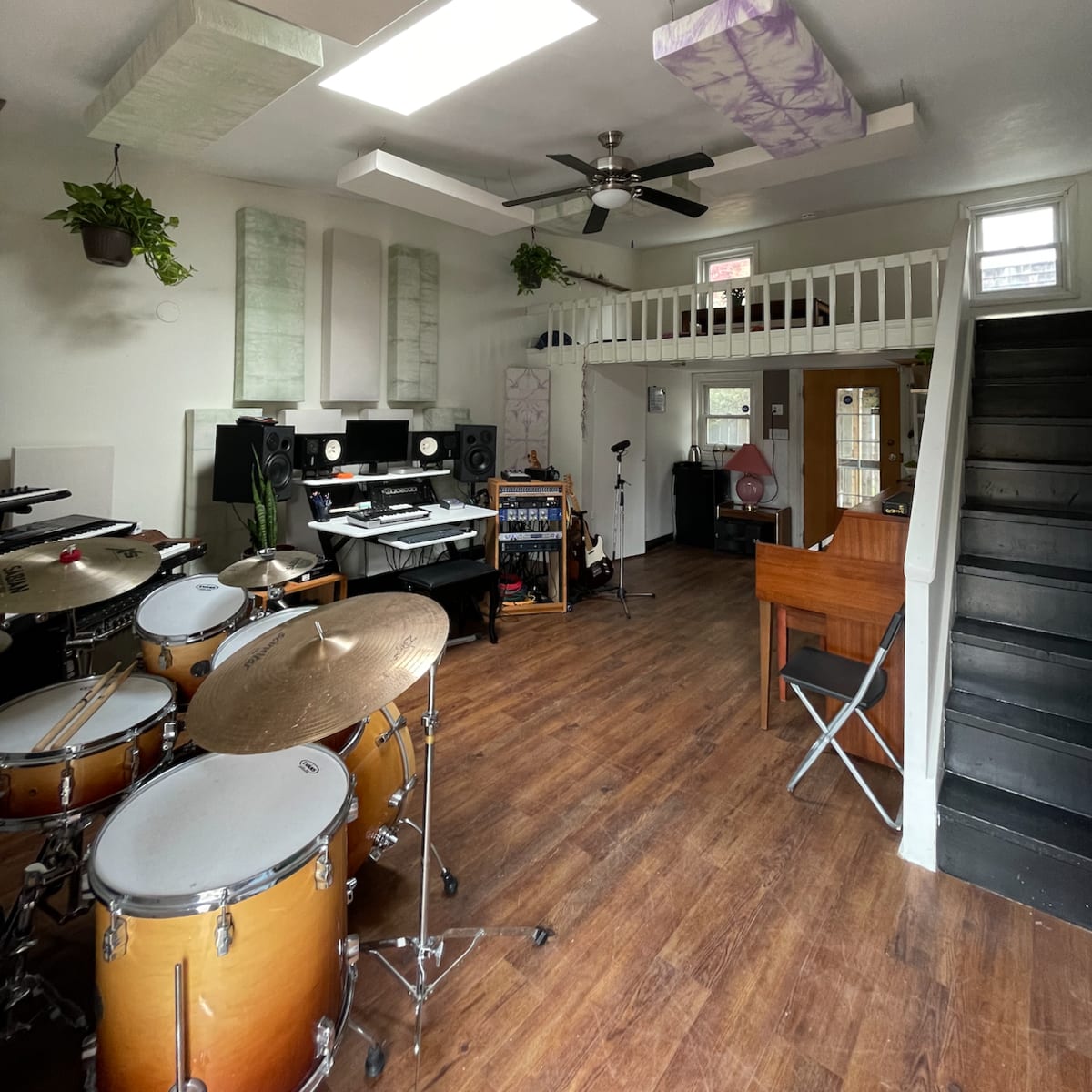 A spacious music studio featuring a drum set, a computer workstation, and a staircase leading to an upper level, with various soundproofing panels and plants decorating the space.