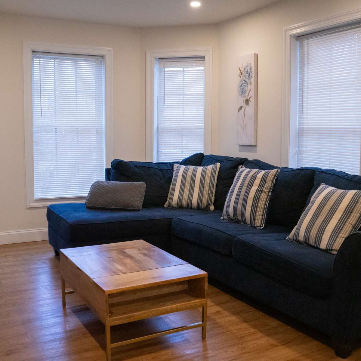 A living room featuring a dark blue sectional sofa with striped cushions, a wooden coffee table, and large windows with blinds.